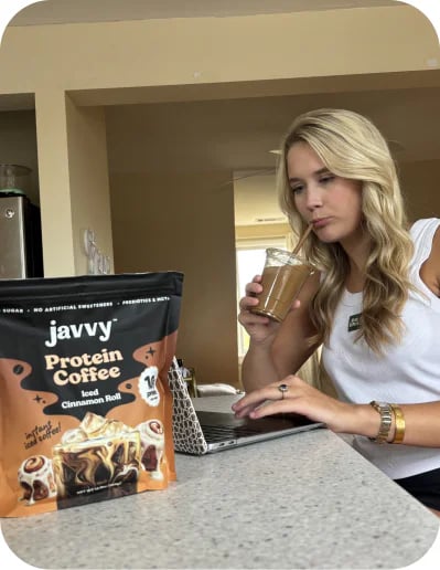 Customer sipping an iced coffee at her laptop with a bag of Iced Cinnamon Roll Protein Coffee