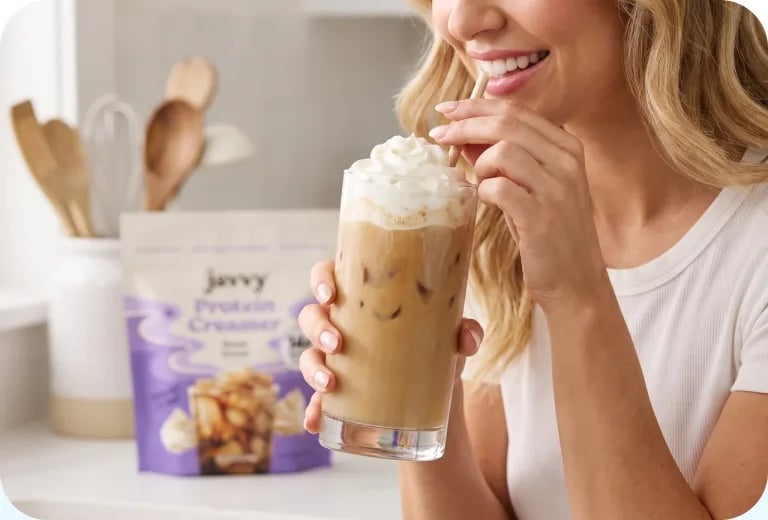 Woman sipping an iced coffee with whipped cream next to a bag of Protein Creamer